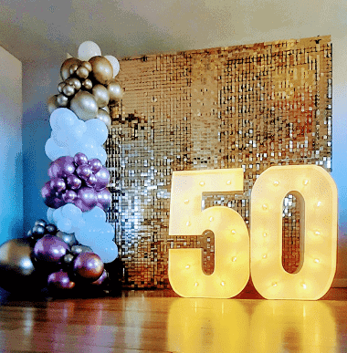 decoración cumpleaños 50 con shimmer walls dorado, guirnalda o arco de globos y números rellenos de luces cinco y cero alusivo a celebración cincuenta años